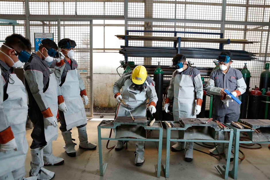Group of workers in full protective suits and masks observing a trainee welding at a metal workbench in a shop. The scene shows safety gear and welding torches nearby.