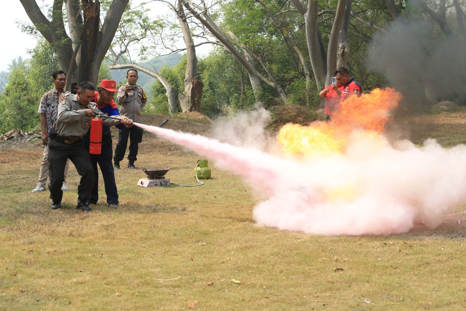 Outdoor fire-safety training: a man aims a fire extinguisher at a small blaze while others look on as orange flames and pink smoke erupt from the flame.