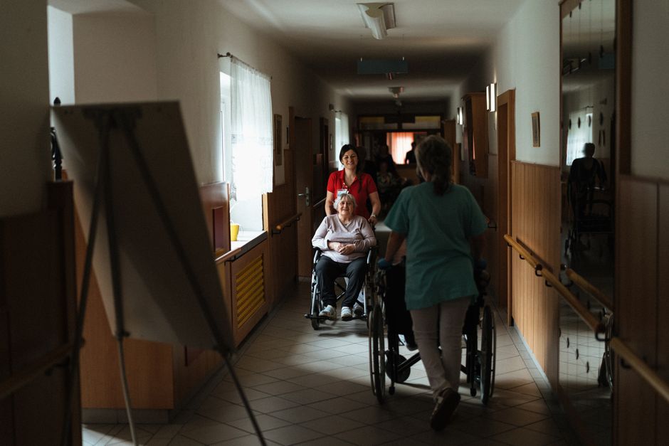 Nurse in a red top pushes an elderly woman in a wheelchair down a sunlit care-facility corridor.