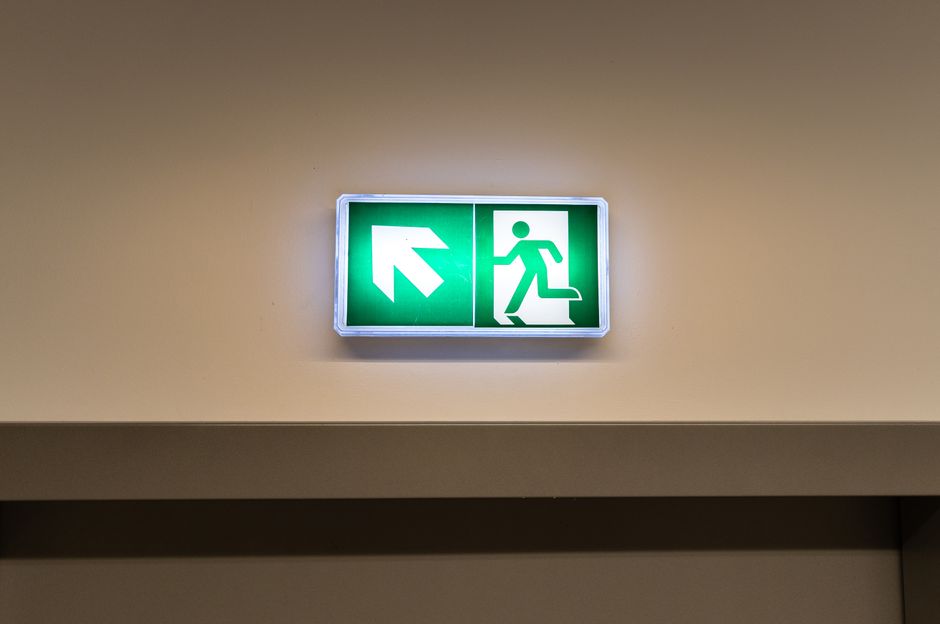 Illuminated green emergency exit sign showing a running figure and an arrow, mounted on a beige wall.