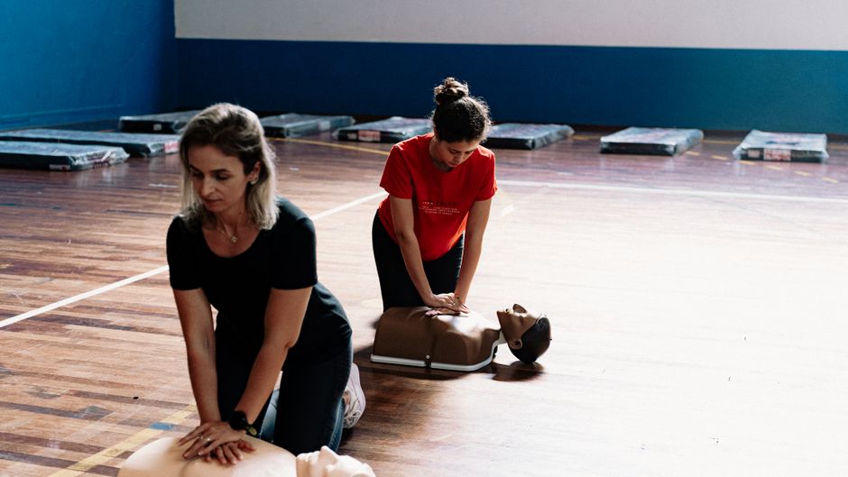 Two people performing CPR on practice mannequins in a gym setting on a wooden floor.