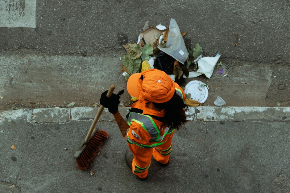 Street cleaner in bright orange uniform sweeping debris into a pile on the curb.