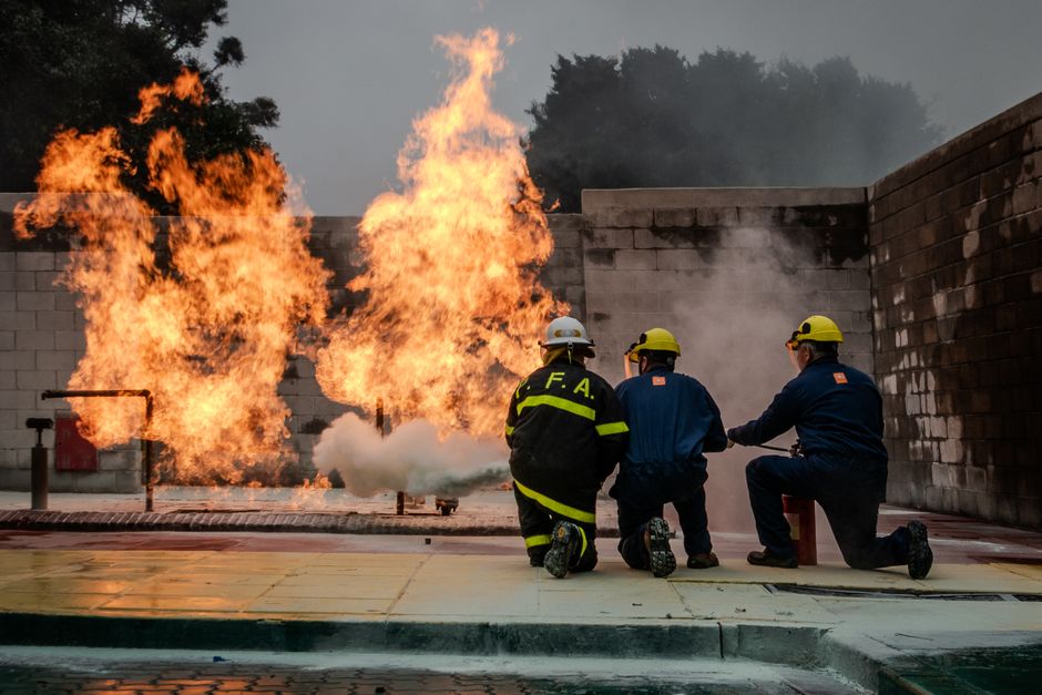 Firefighters in full gear kneel and monitor a large training flame in a fenced outdoor area with brick walls, practicing fire suppression.