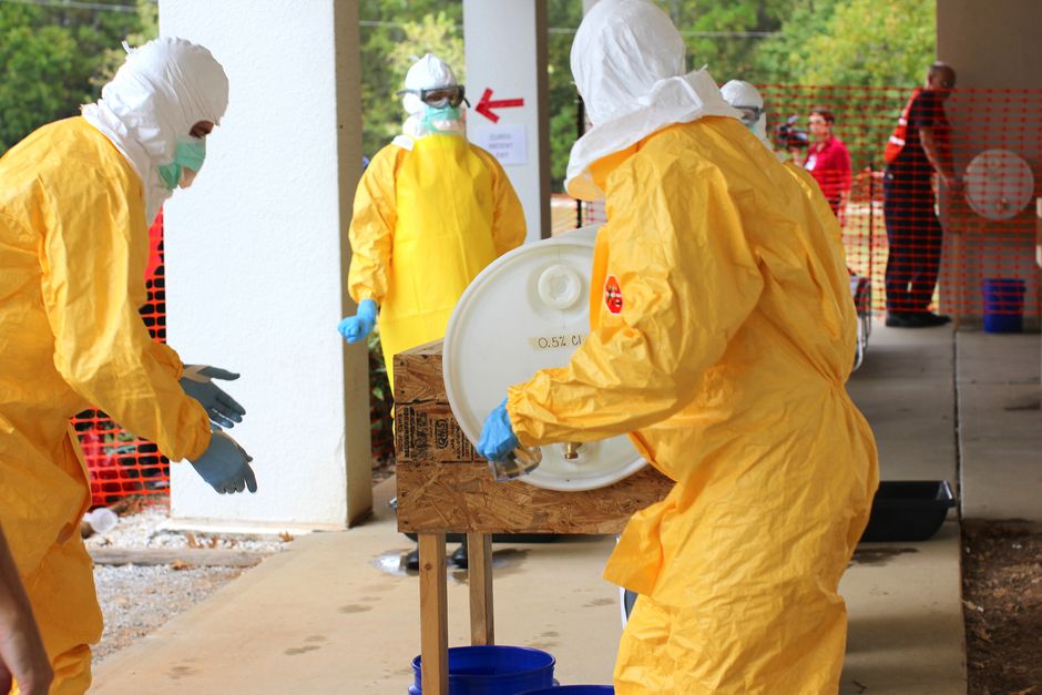 Hazmat-suited workers in yellow protective suits handling a large drum on a wooden stand during a chemical spill response under a covered outdoor area.