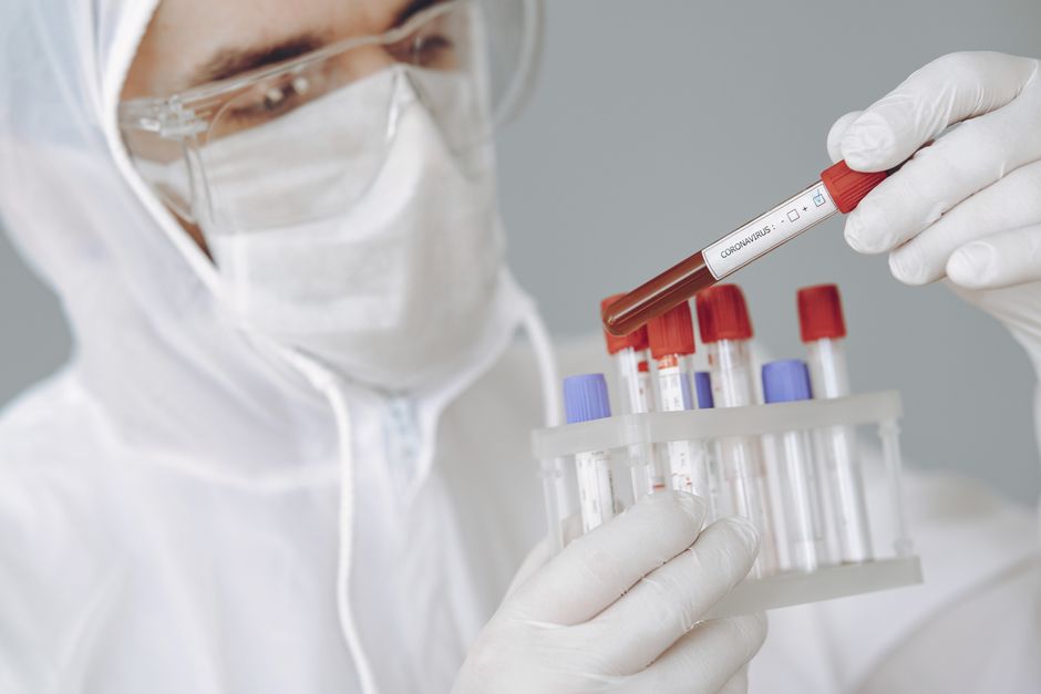 Laboratory technician in PPE holds a blood sample labeled 'CORONAVIRUS' in a test tube rack for testing.