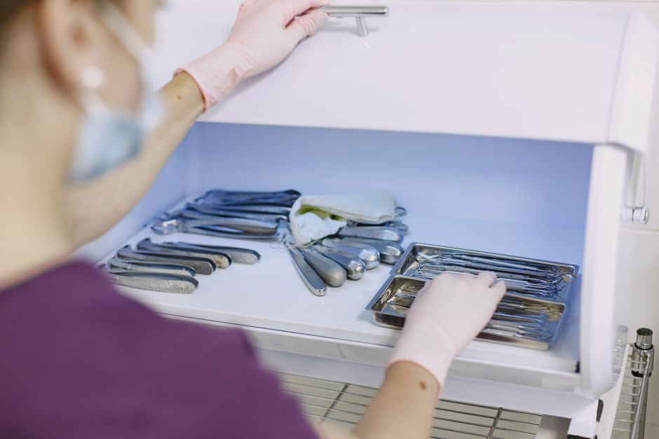 Healthcare worker in scrubs with gloves and a mask places sterilized instruments into a medical cabinet drawer.