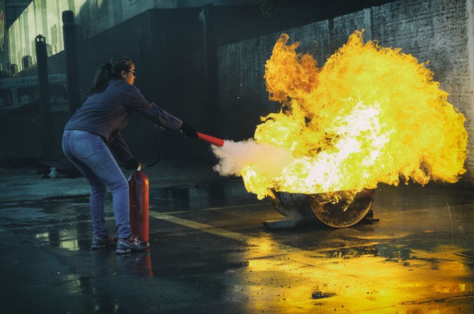 Person in jeans uses a red fire extinguisher to spray a large indoor flame. (Outdoor alley scene)