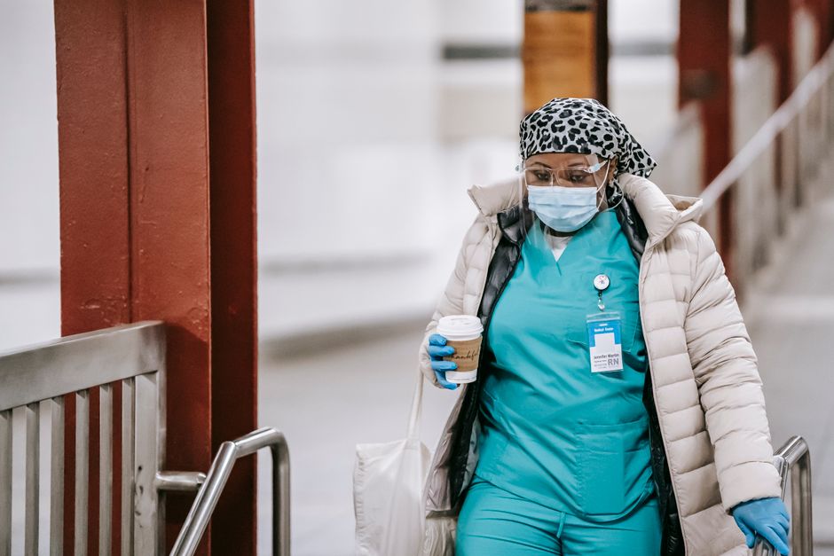 Nurse in teal scrubs with mask, gloves, and a leopard-print cap carrying a coffee cup in a hospital corridor.