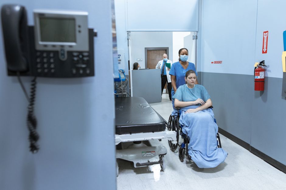 Nurse in blue scrubs pushes a patient in a wheelchair down a hospital hallway, both wearing masks.