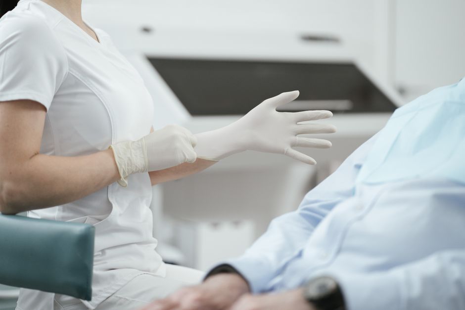 Nurse in white scrubs with gloves reaching toward a patient in a hospital bed, ready to assist.