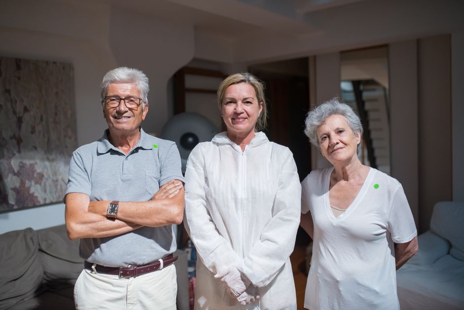 Three adults stand together indoors, smiling at the camera: a man in a polo on the left, a woman in a white coat in the center, and a woman in a white shirt on the right.